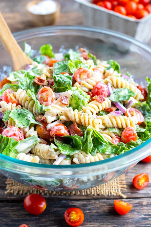 A glass Pyrex bowl full of a BLT pasta salad being mixed together with a wooden spoon.