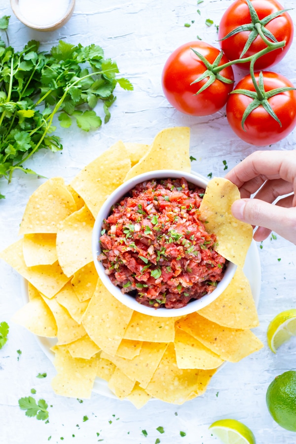 A hand dipping a tortilla chip into a bowl full of restaurant style tomato salsa with fresh tomatoes and cilantro next to it.