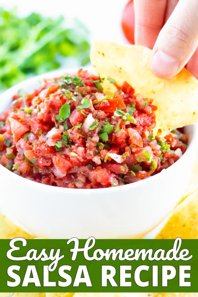 A thick and chunky salsa recipe in a white bowl and a hand dipping a tortilla chip into it.