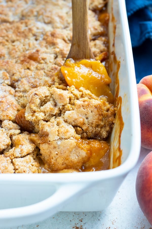 Tender peaches and a buttery topping are being served out of a baking dish for a southern peach cobbler dessert.