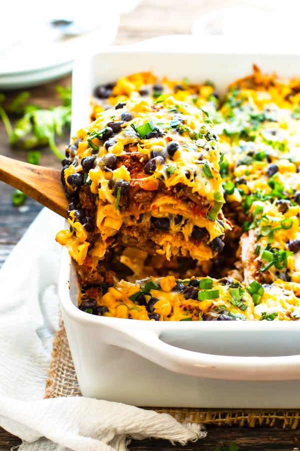 A piece of a Mexican lasagna recipe being pulled from a white casserole dish with a white spoon.