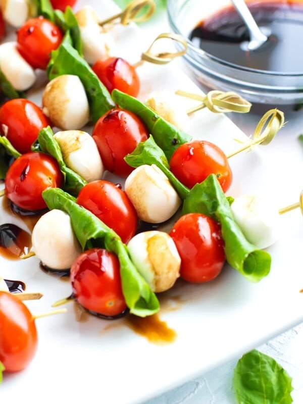 Caprese salad skewers on a white serving platter with a clear bowl full of balsamic glaze.