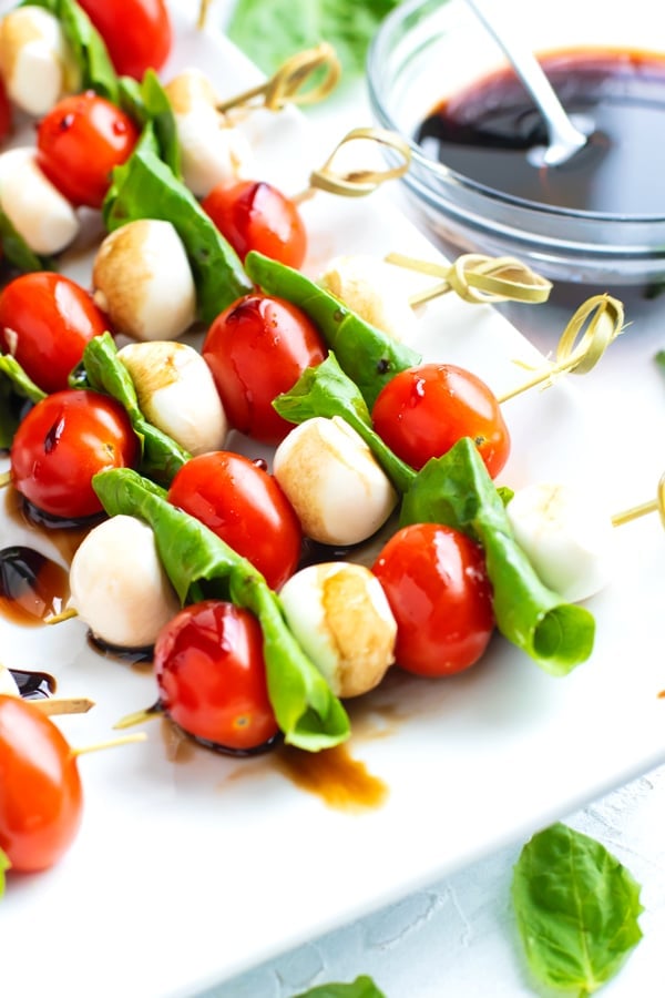 Caprese salad skewers on a white serving platter with a clear bowl full of balsamic glaze.