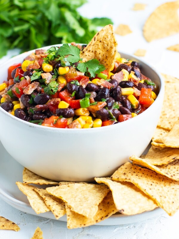 Black bean dip recipe with corn, green onions, and salsa in a white bowl with corn tortilla chips.