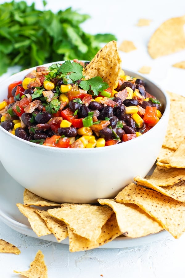 Black bean dip recipe with corn, green onions, and salsa in a white bowl with corn tortilla chips.