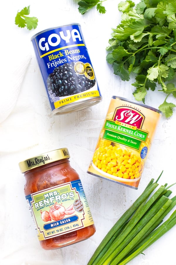 A can of black beans, a can of kernel corn, mild salsa, cilantro, and green onions on a white background.