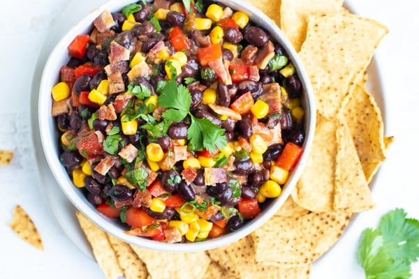 Black bean bacon dip for a party appetizer in a white bowl surrounded by corn tortilla chips.