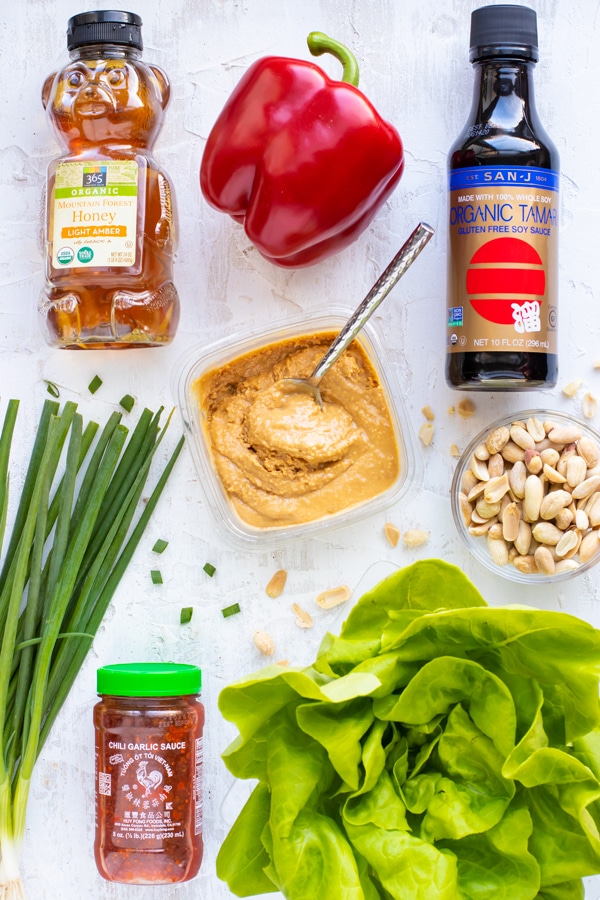A bottle of honey, a red bell pepper, gluten-free soy sauce, smooth peanut butter, Sriracha sauce, roasted peanuts, and Bibb lettuce on a white table as the ingredients for an Asian lettuce wraps.