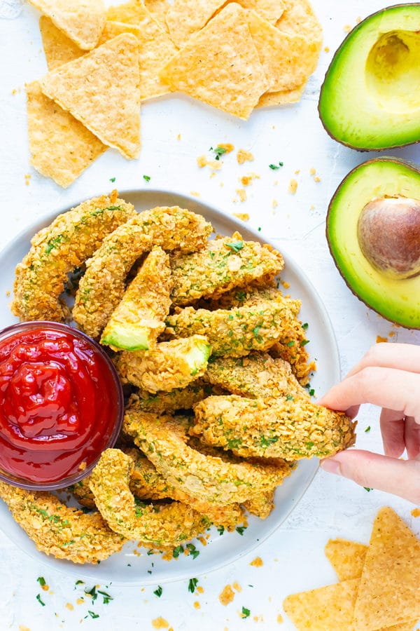 A hand that is grabbing an avocado fry from a white plate full of avocado fries with tortilla chips surrounding it.