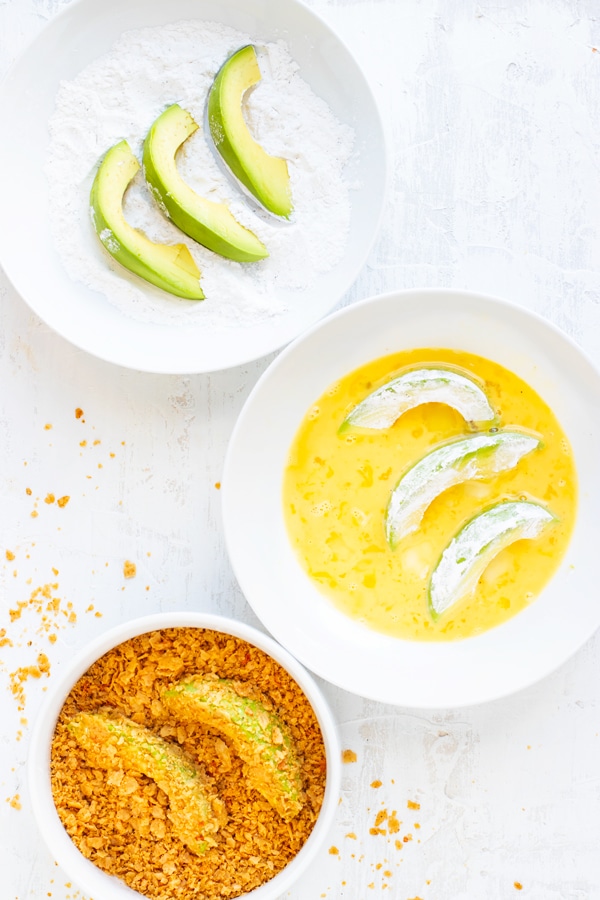 Three white bowls with avocado slices in starch, avocado slices in an egg wash, and avocado fries in a tortilla chip crust for an avocado fries recipe.