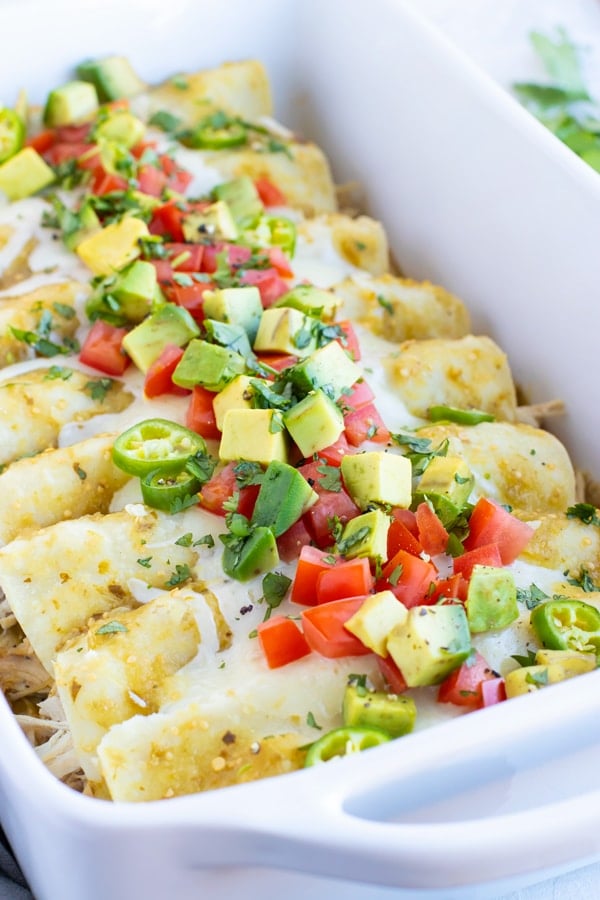 Chicken Enchiladas Verdes recipe in a white baking dish with avocado, tomatoes, and cilantro on top.