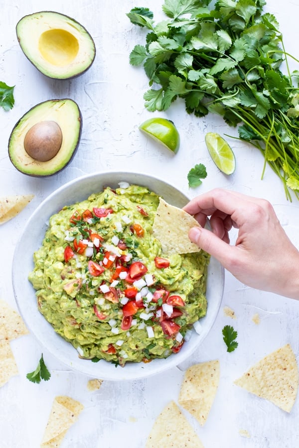 A hand dipping a corn tortilla chip into a big bowl full of a simple guacamole with tomatoes and onions.