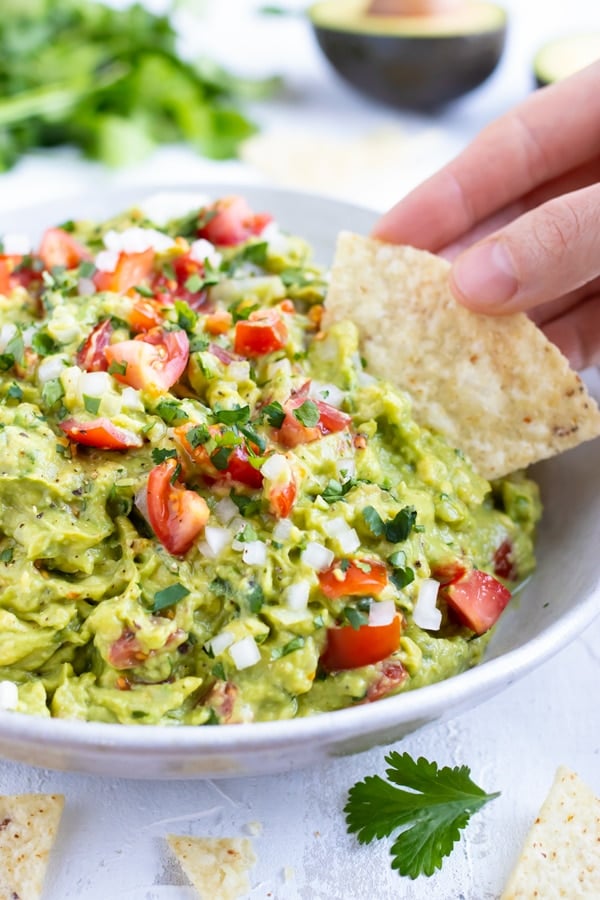 A tortilla chip being dipped into a healthy guacamole dip with tomatoes, onions, and cilantro.