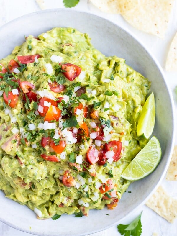 A white bowl full of a simple guacamole recipe with limes, tomatoes, and onions to garnish.