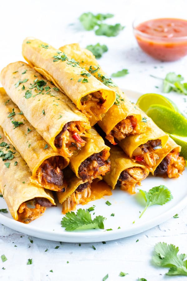 A stack of baked chicken taquitos with refried beans and cheese on a white plate for a Cinco de Mayo party.