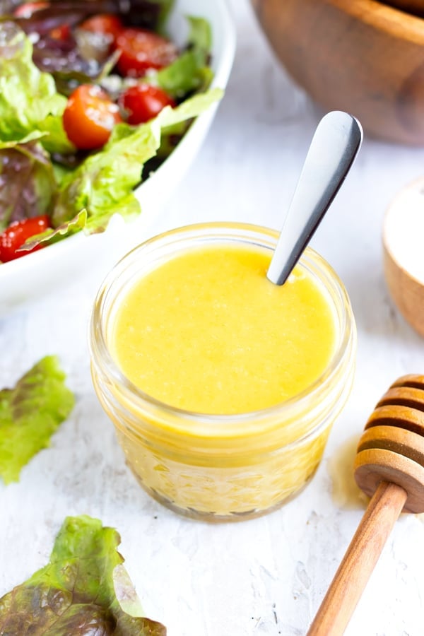 A jar full of honey mustard dressing with a silver spoon next to two wooden salad bowls.