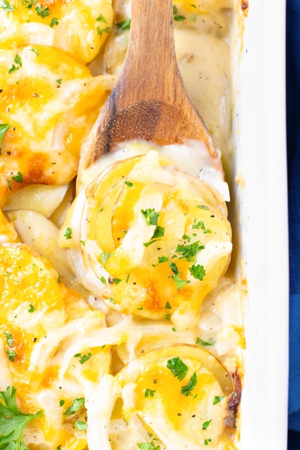 A close-up of a wooden spoon scooping out a serving of scalloped potatoes with cheese.