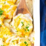 A wooden serving spoon picking up a portion of scalloped potatoes from a baking pan.