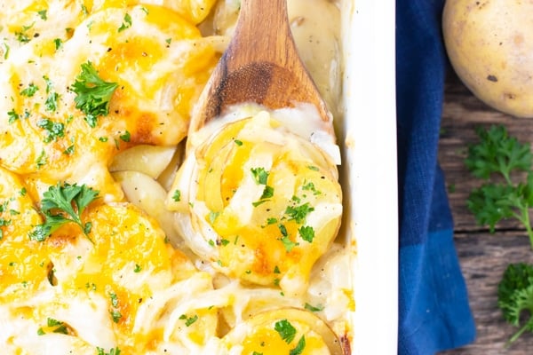 A wooden serving spoon picking up a portion of scalloped potatoes from a baking pan.