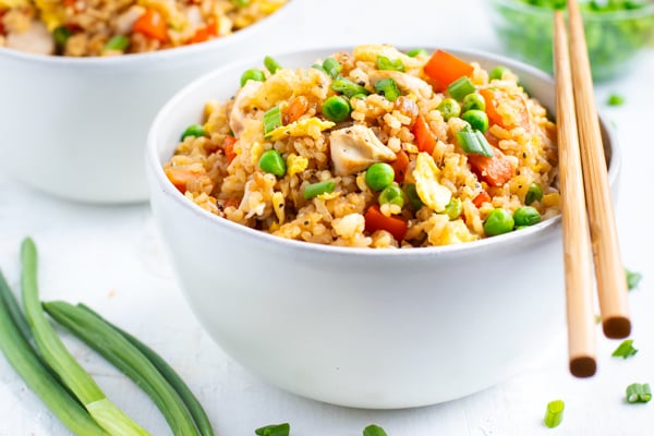 A white bowl full of a healthy fried rice recipe with carrots and peas next to green onions.