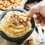 A hand dipping a pretzel into a bowl of hummus made with a gluten-free lentil recipe.