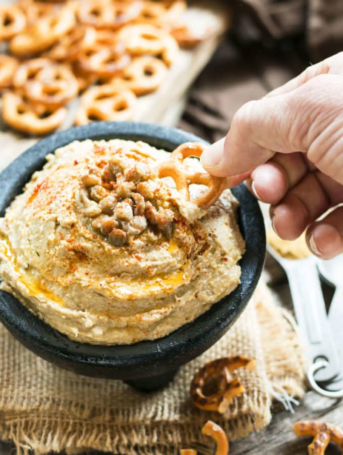 A hand dipping a pretzel into a bowl of hummus made with a gluten-free lentil recipe.