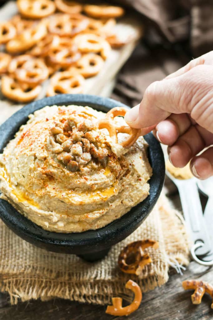 A hand dipping a pretzel into a bowl of hummus made with a gluten-free lentil recipe.