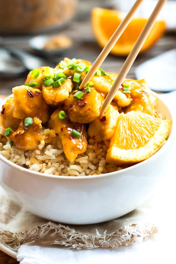 A pair of chopsticks attempting to pick up a piece of gluten-free Chinese orange chicken in a bowl.