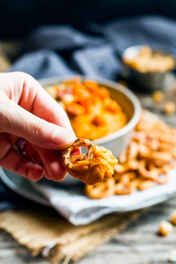 A hand holding a pretzel dipped in gluten-free sweet potato hummus for a healthy snack.