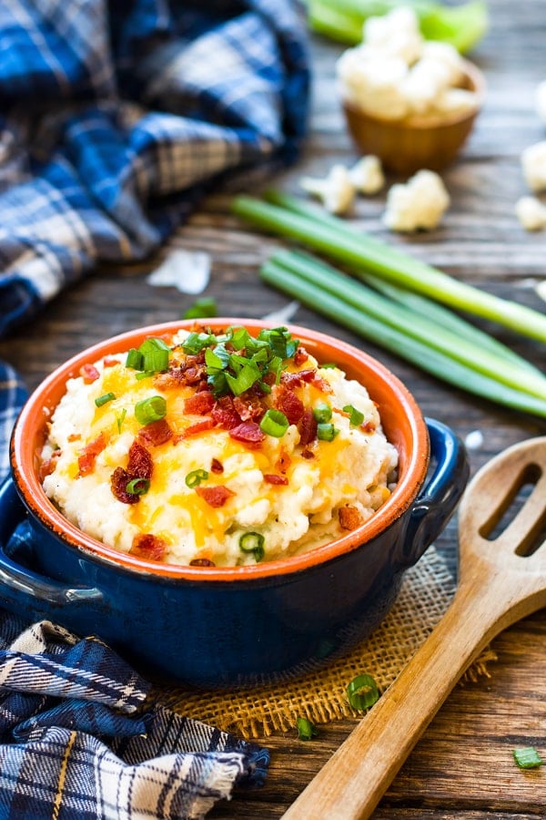 A bowl of a gluten-free cauliflower mashed potato recipe on a table topped with green onions and bacon.
