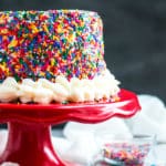 The best Funfetti Cake with vanilla on a red cake plate surrounded by rainbow sprinkles.