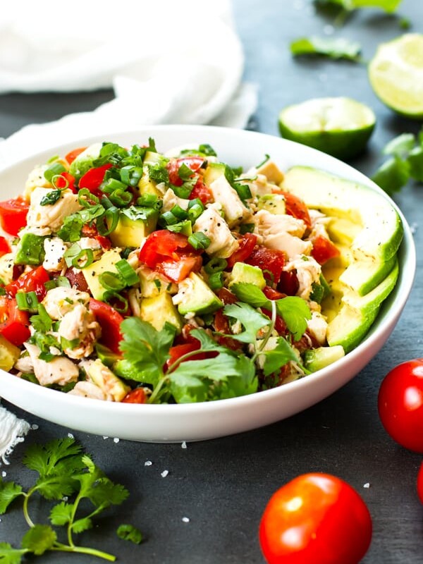 A Tomato and Avocado Chicken Salad in a white bowl with limes and cilantro on the side.