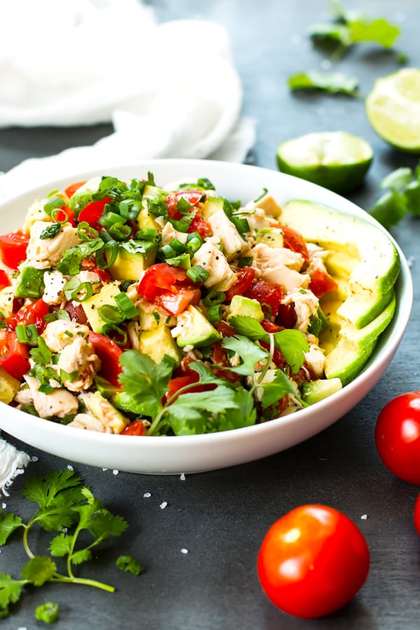 A Tomato and Avocado Chicken Salad in a white bowl with limes and cilantro on the side.