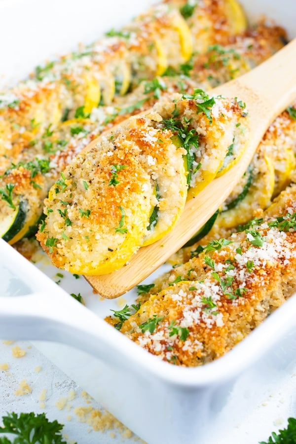 A serving spoon scooping out baked squash and zucchini casserole from a white casserole dish.