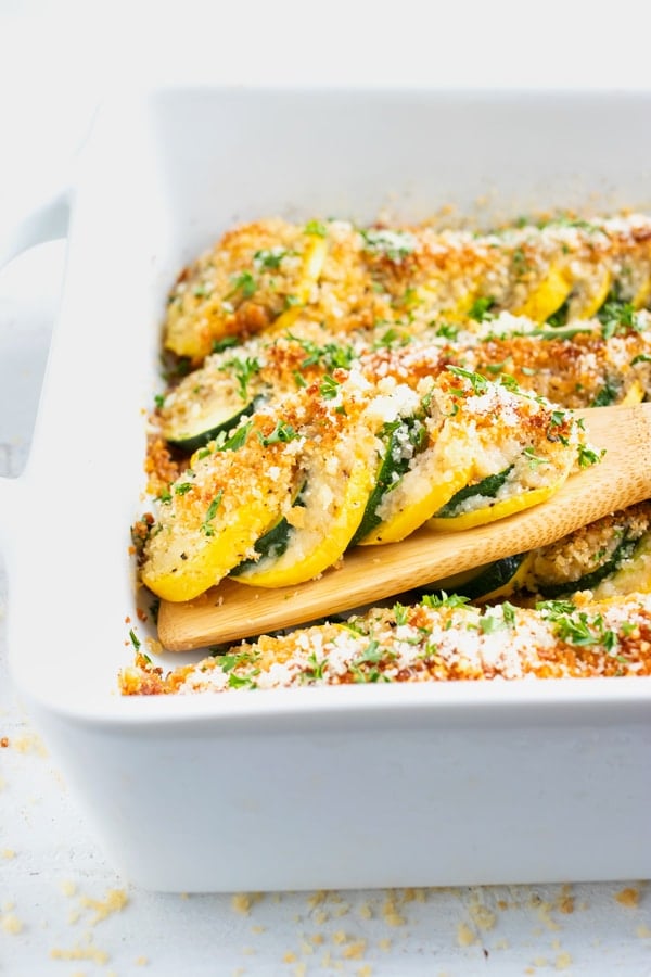 A wooden spoon picking up baked zucchini and summer squash from a white baking dish.