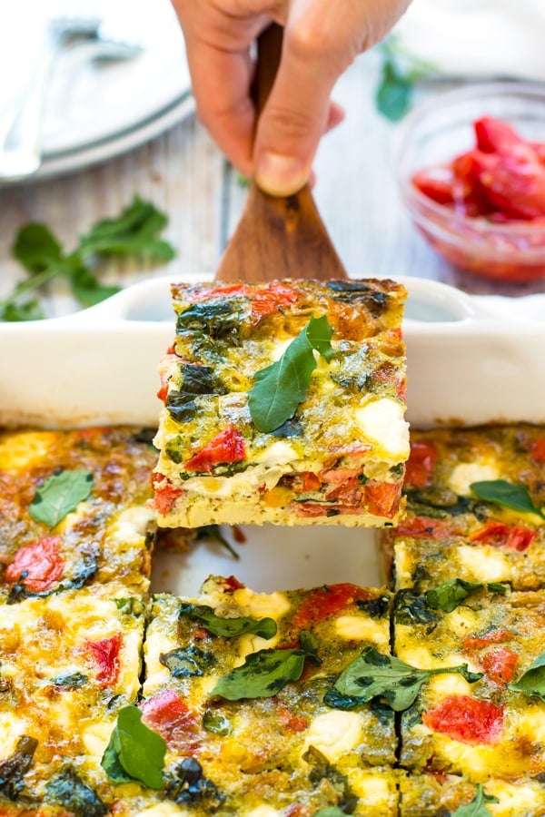 A hand taking a serving of a baked frittata recipe out of a casserole dish for brunch.