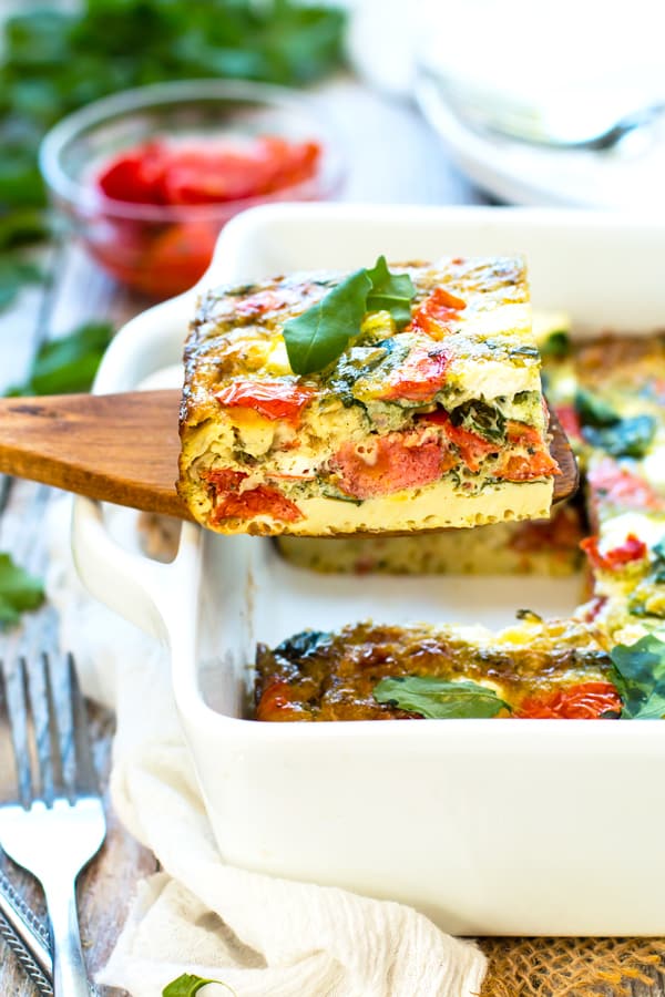 A wooden serving spoon holding a serving of a Baked Frittata with Pesto, Tomatoes, and Goat Cheese.