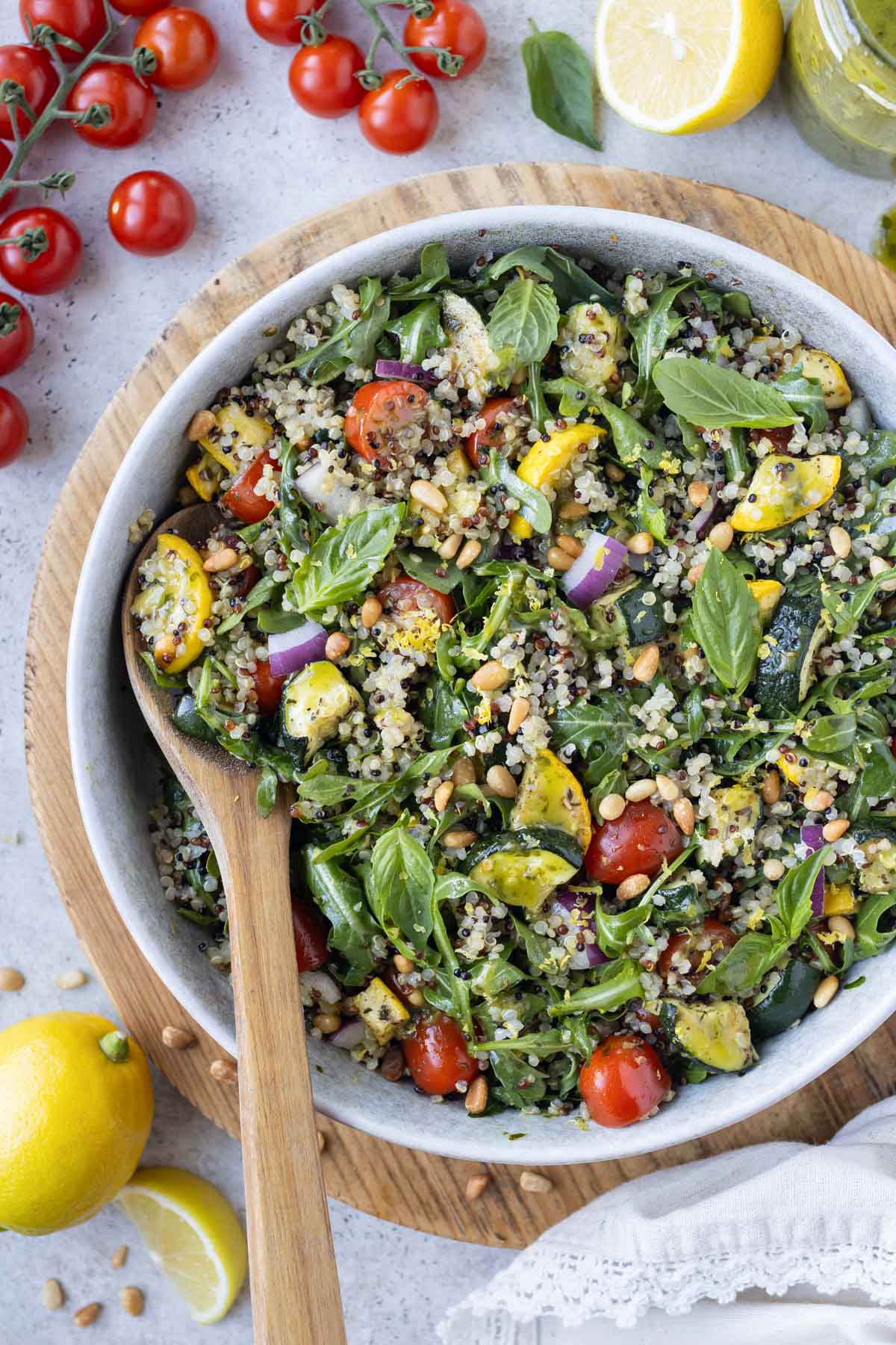 A wooden spoon stirs a large bowl of summer quinoa salad with roasted veggies.