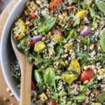 A summer quinoa salad with a lemon vinaigrette, summer squash, and tomatoes is served in a white bowl with a wooden spoon.