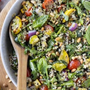 A summer quinoa salad with a lemon vinaigrette, summer squash, and tomatoes is served in a white bowl with a wooden spoon.