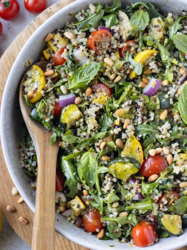 A summer quinoa salad with a lemon vinaigrette, summer squash, and tomatoes is served in a white bowl with a wooden spoon.