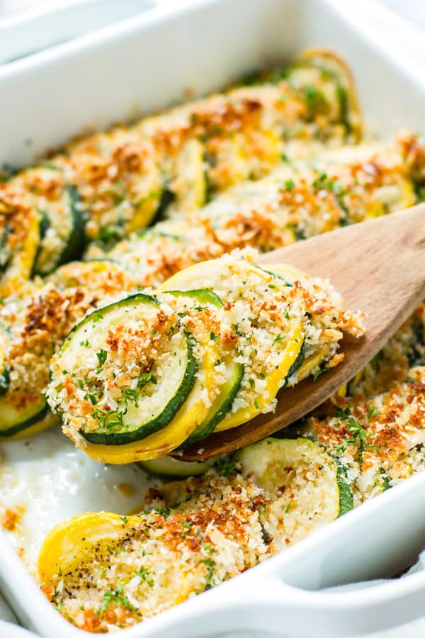 A serving spoon scooping out baked squash and zucchini casserole from a white casserole dish.