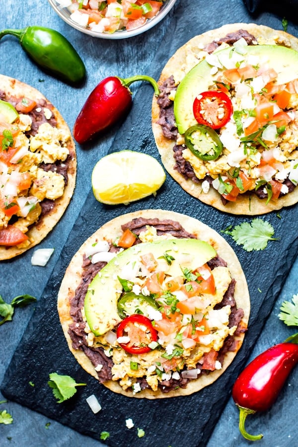 Two breakfast tostadas on a blue slab with peppers on the side.