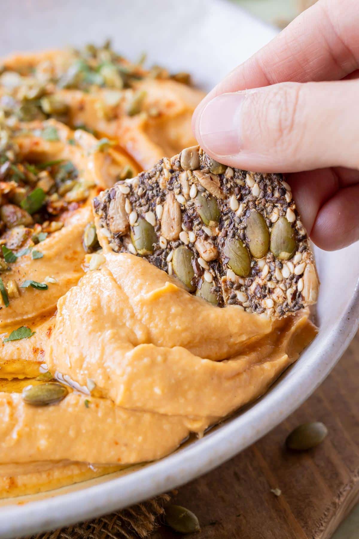 A flaxseed cracker scoops up some pumpkin hummus from a dip bowl.