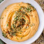 A savory pumpkin hummus recipe is in a white bowl with roasted pumpkin seeds on top.