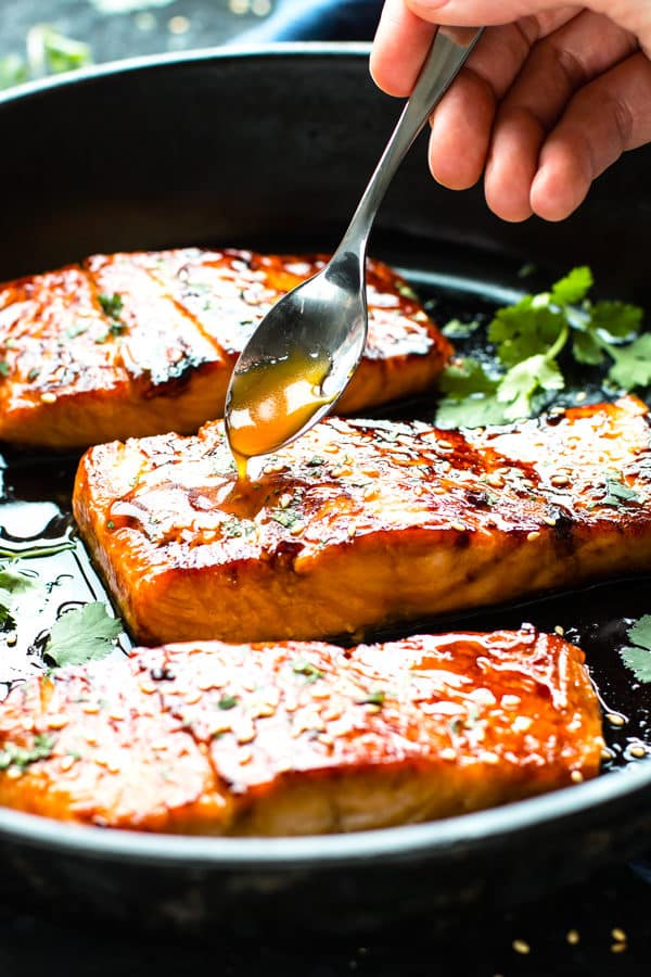 Honey sriracha sauce being drizzled on a salmon fillet in a black pan.