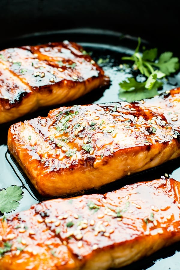Three honey glazed salmon fillets in a black pan with fresh herbs.