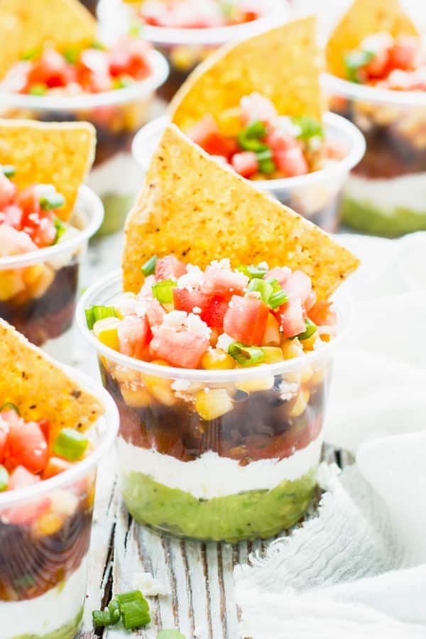 A collection of plastic cups containing healthy 7-layer dips with tortilla chips as a topping.