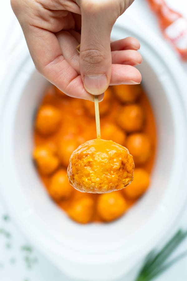 A hand holding a chicken meatball covered in buffalo wing sauce over a Crock-pot.