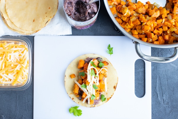 Assembling a vegetarian taco on a cutting board with sweet potatoes, refried black beans, and cheese.